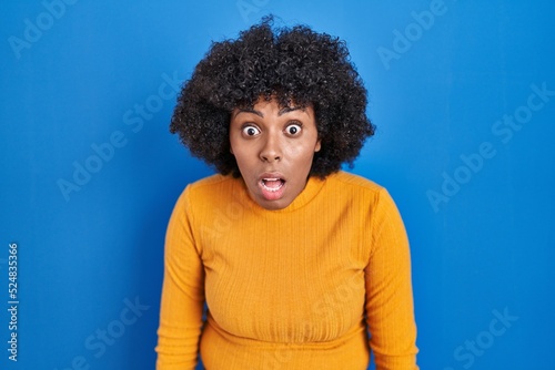 Black woman with curly hair standing over blue background afraid and shocked with surprise and amazed expression, fear and excited face.