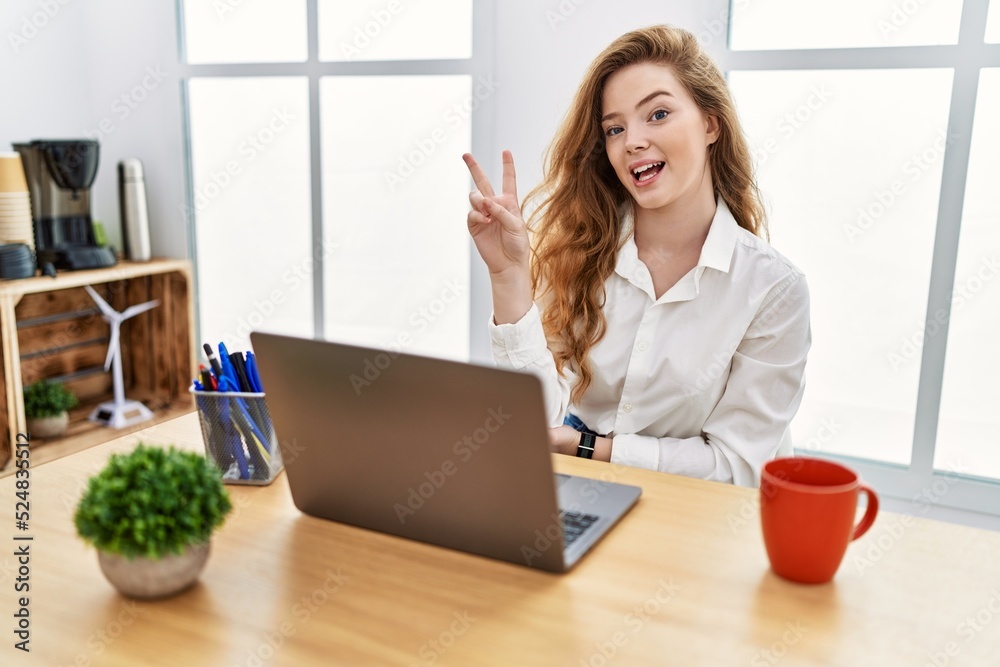 Young caucasian woman working at the office using computer laptop smiling with happy face winking at the camera doing victory sign with fingers. number two. Young caucasian woman working at the office using computer laptop smiling with happy face winking at the camera doing victory sign with fingers. number two.