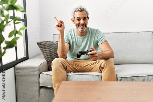Middle age hispanic man playing video game sitting on the sofa smiling happy pointing with hand and finger to the side