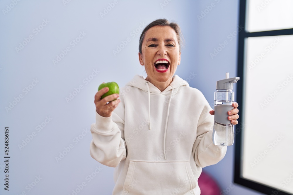 Middle age woman smiling confident holding apple and bottle of water at sport center Middle age woman smiling confident holding apple and bottle of water at sport center