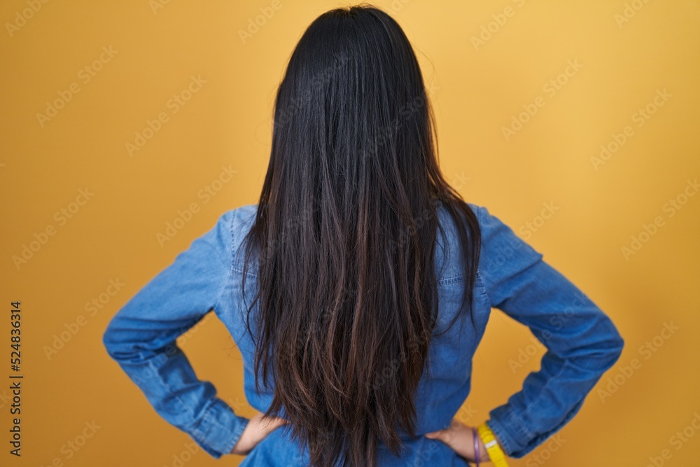 Fototapeta premium Young asian woman standing over yellow background standing backwards looking away with arms on body