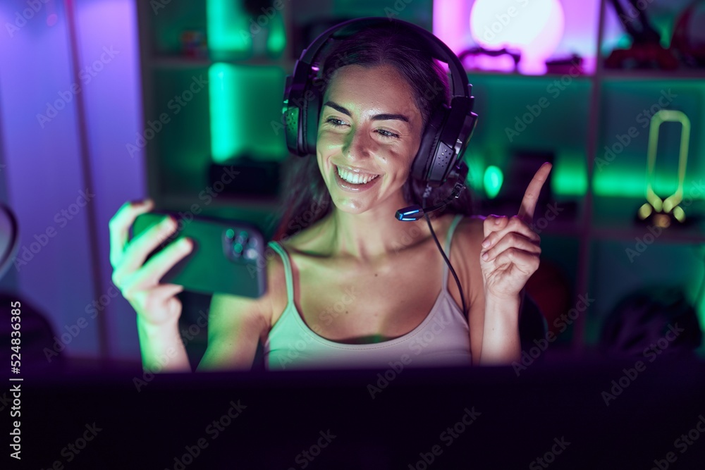 Young brunette woman playing video games with smartphone smiling happy pointing with hand and finger to the side
