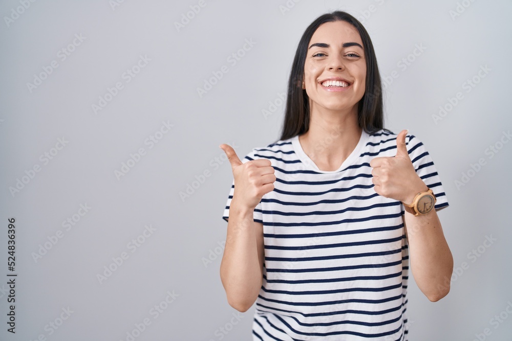 Young brunette woman wearing striped t shirt success sign doing positive gesture with hand, thumbs up smiling and happy. cheerful expression and winner gesture. Young brunette woman wearing striped t shirt success sign doing positive gesture with hand, thumbs up smiling and happy. cheerful expression and winner gesture.