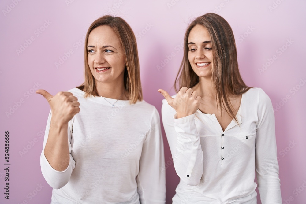 Middle age mother and young daughter standing over pink background smiling with happy face looking and pointing to the side with thumb up. Middle age mother and young daughter standing over pink background smiling with happy face looking and pointing to the side with thumb up.