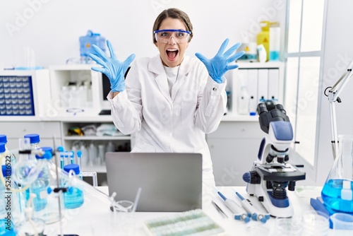 Young hispanic woman wearing scientist uniform working at laboratory celebrating victory with happy smile and winner expression with raised hands