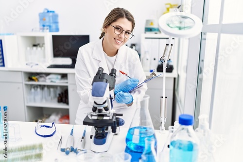 Young hispanic woman wearing scientist uniform using microscope at laboratory