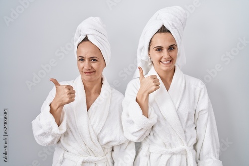 Middle age woman and daughter wearing white bathrobe and towel doing happy thumbs up gesture with hand. approving expression looking at the camera showing success.