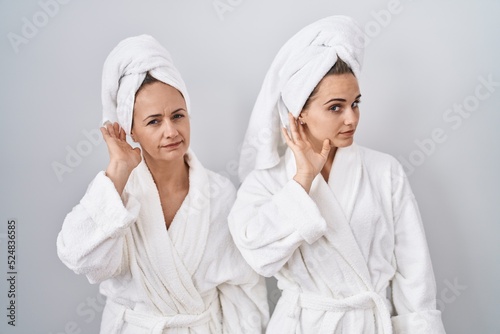 Middle age woman and daughter wearing white bathrobe and towel smiling with hand over ear listening an hearing to rumor or gossip. deafness concept.
