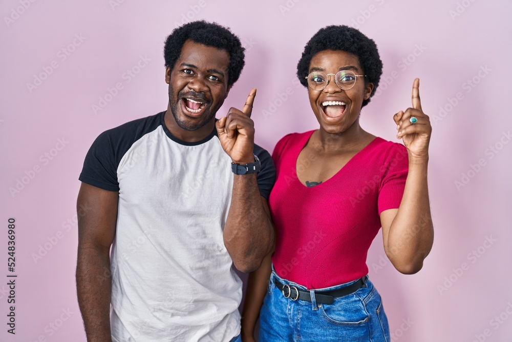 Young african american couple standing over pink background pointing finger up with successful idea. exited and happy. number one. Young african american couple standing over pink background pointing finger up with successful idea. exited and happy. number one.