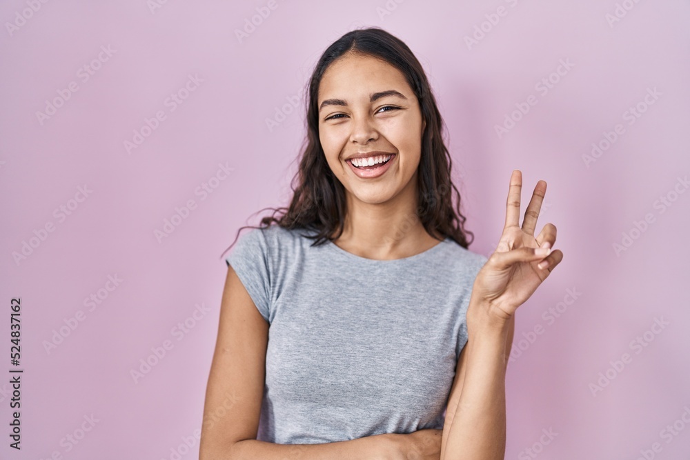 Young brazilian woman wearing casual t shirt over pink background smiling with happy face winking at the camera doing victory sign. number two. Young brazilian woman wearing casual t shirt over pink background smiling with happy face winking at the camera doing victory sign. number two.