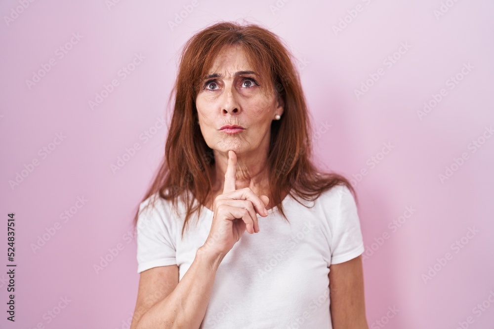Middle age woman standing over pink background thinking concentrated about doubt with finger on chin and looking up wondering Middle age woman standing over pink background thinking concentrated about doubt with finger on chin and looking up wondering