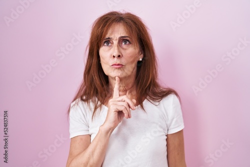 Middle age woman standing over pink background thinking concentrated about doubt with finger on chin and looking up wondering