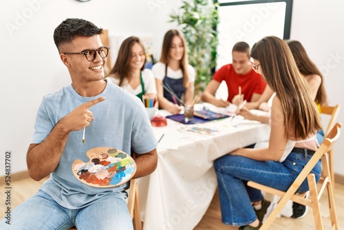 Young artist man drawing at art studio pointing thumb up to the side smiling happy with open mouth