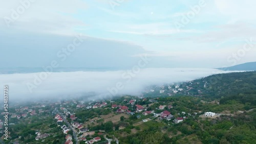 Flying above the fog over Pécs Hungary in the morning with mountain in background over village at dawn with cloudy sky while rotating camera 4K