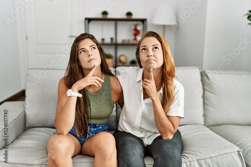 Mother and daughter together sitting on the sofa at home thinking concentrated about doubt with finger on chin and looking up wondering