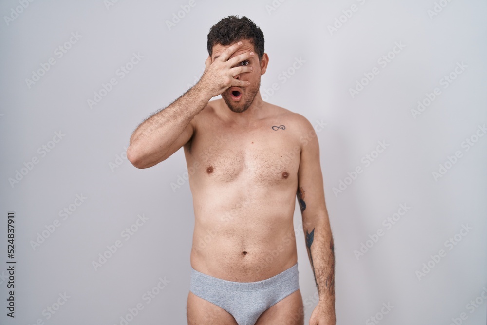 Young hispanic man standing shirtless wearing underware peeking in shock covering face and eyes with hand, looking through fingers with embarrassed expression.