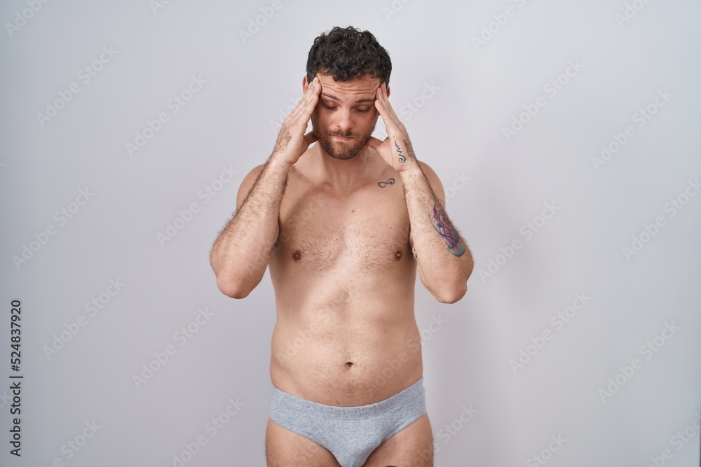 Young hispanic man standing shirtless wearing underware with hand on head, headache because stress. suffering migraine.