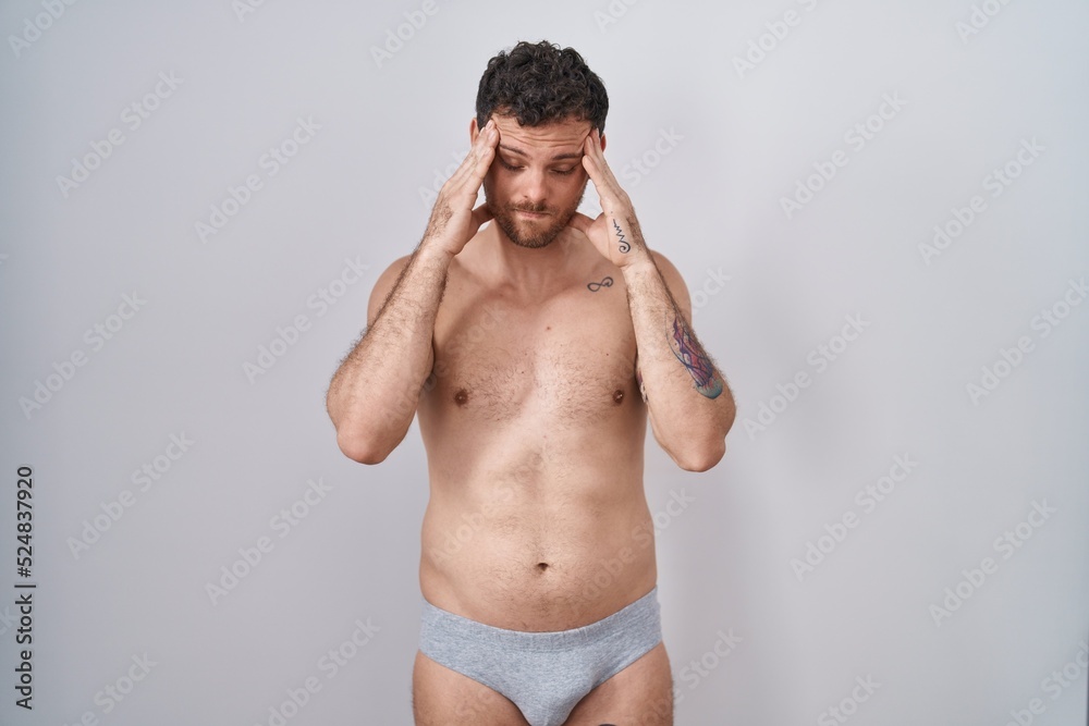Fototapeta premium Young hispanic man standing shirtless wearing underware with hand on head, headache because stress. suffering migraine.