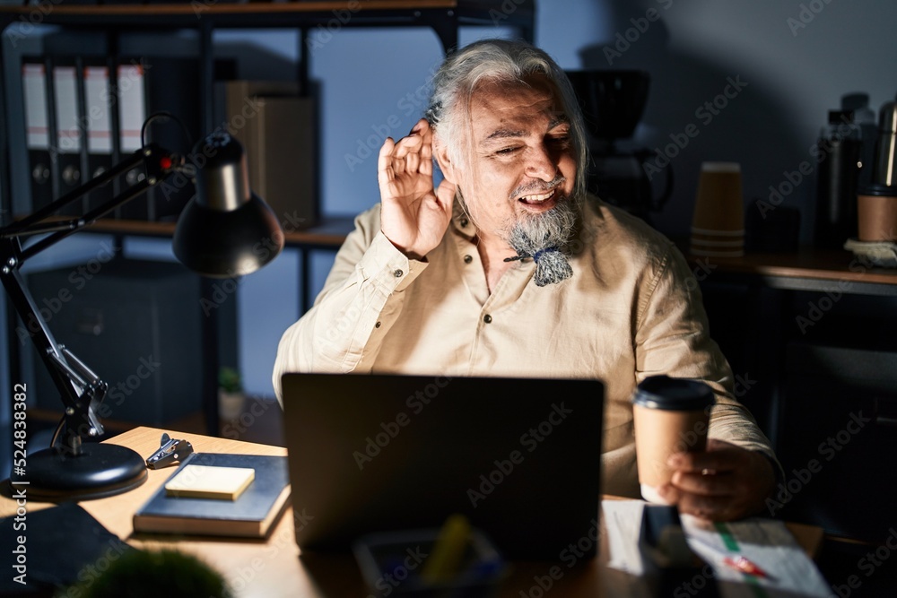 Middle age man with grey hair working at the office at night smiling with hand over ear listening an hearing to rumor or gossip. deafness concept. Middle age man with grey hair working at the office at night smiling with hand over ear listening an hearing to rumor or gossip. deafness concept.