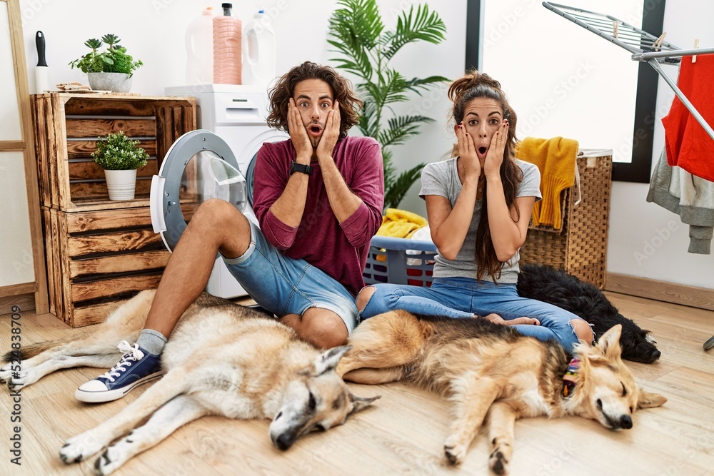 Young hispanic couple doing laundry with dogs afraid and shocked, surprise and amazed expression with hands on face