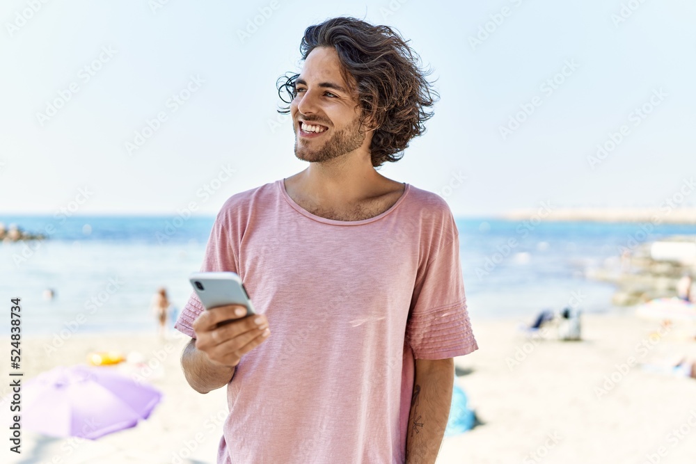 Young hispanic man smiling happy using smartphone at the beach.