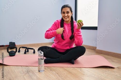 Young hispanic woman sitting on yoga mat eating apple pointing finger to one self smiling happy and proud