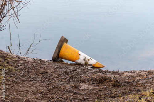 A tatty discarded traffic cone lying on its side in some shallow water next to a muddy bank.
