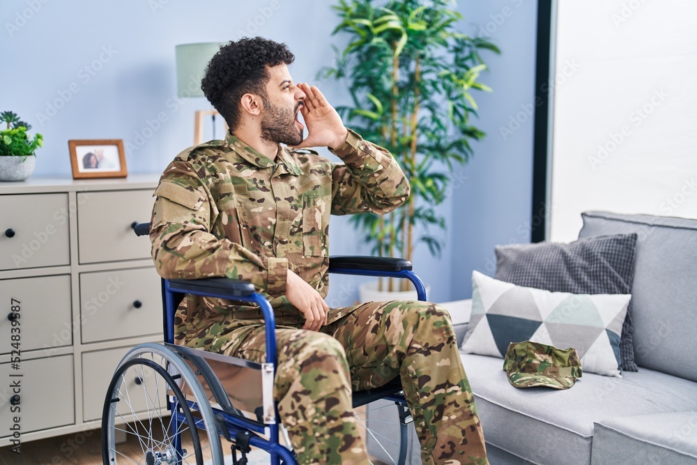 Arab man wearing camouflage army uniform sitting on wheelchair shouting and screaming loud to side with hand on mouth. communication concept.