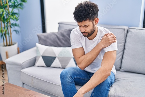 Young arab man suffering for shoulder injury sitting on sofa at home