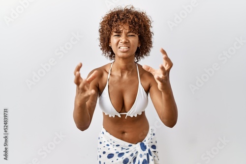 Young african american woman with curly hair wearing bikini shouting frustrated with rage, hands trying to strangle, yelling mad
