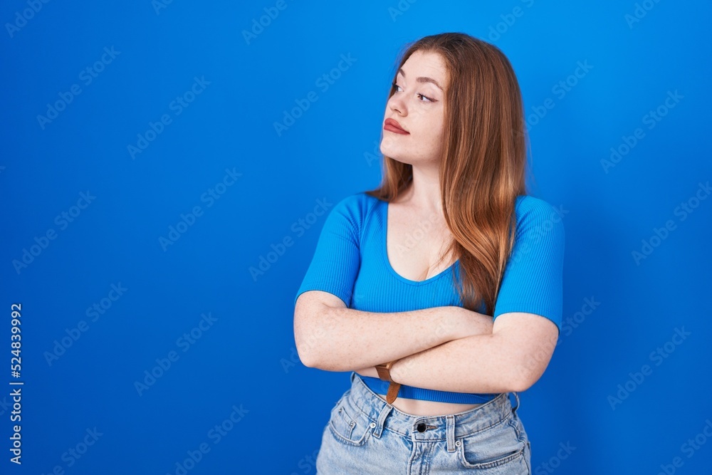 Redhead woman standing over blue background looking to the side with arms crossed convinced and confident Redhead woman standing over blue background looking to the side with arms crossed convinced and confident