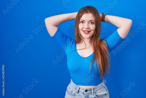 Redhead woman standing over blue background relaxing and stretching, arms and hands behind head and neck smiling happy