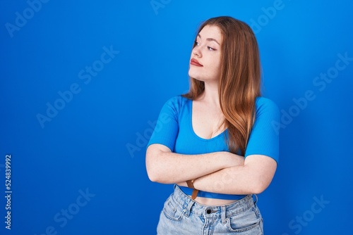 Redhead woman standing over blue background looking to the side with arms crossed convinced and confident