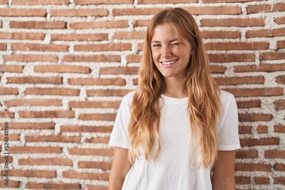 Young caucasian woman standing over bricks wall winking looking at the camera with sexy expression, cheerful and happy face.