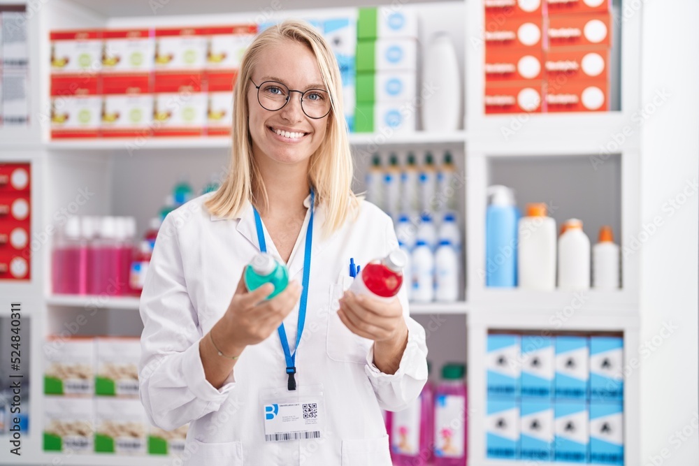 Young blonde woman pharmacist smiling confident holding medication bottles at pharmacy