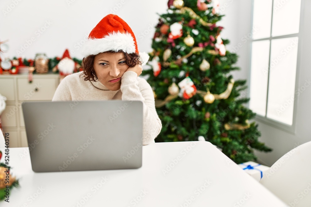 Middle age hispanic woman using laptop sitting by christmas tree thinking looking tired and bored with depression problems with crossed arms. Middle age hispanic woman using laptop sitting by christmas tree thinking looking tired and bored with depression problems with crossed arms.