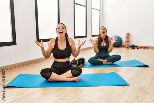 Two hispanic women friends training at the gym sitting on yoga mat with dog celebrating mad and crazy for success with arms raised and closed eyes screaming excited. winner concept