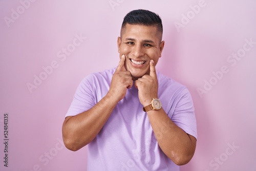 Young hispanic man standing over pink background smiling with open mouth, fingers pointing and forcing cheerful smile