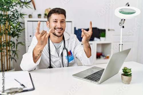 Young doctor working at the clinic using computer laptop shouting with crazy expression doing rock symbol with hands up. music star. heavy concept. © Krakenimages.com