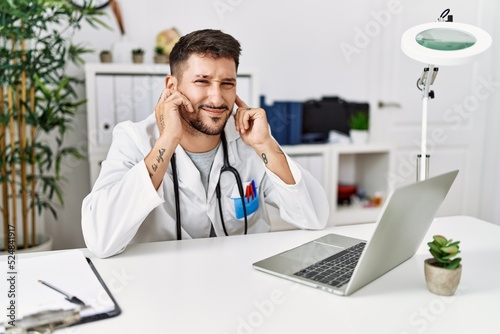 Young doctor working at the clinic using computer laptop covering ears with fingers with annoyed expression for the noise of loud music. deaf concept.