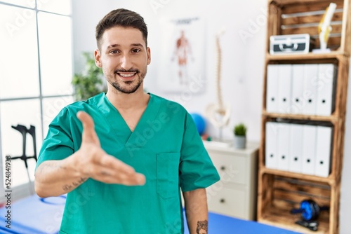 Young physiotherapist man working at pain recovery clinic smiling friendly offering handshake as greeting and welcoming. successful business.