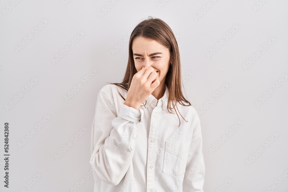 Young caucasian woman standing over isolated background smelling something stinky and disgusting, intolerable smell, holding breath with fingers on nose. bad smell