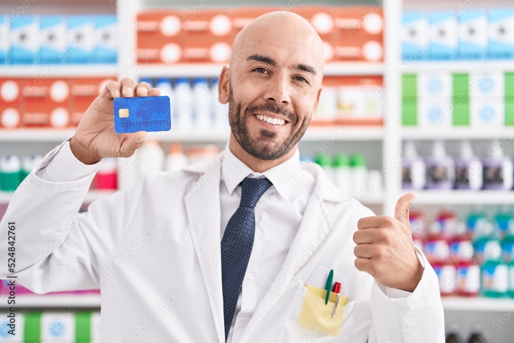 Middle age bald man working at pharmacy drugstore holding credit card smiling happy and positive, thumb up doing excellent and approval sign Middle age bald man working at pharmacy drugstore holding credit card smiling happy and positive, thumb up doing excellent and approval sign
