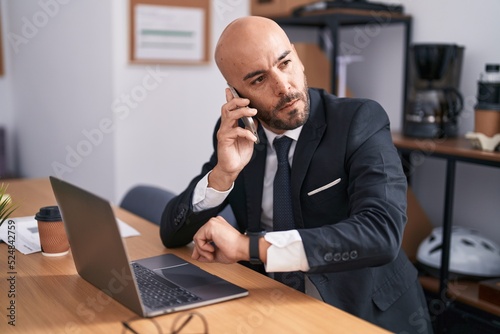 Young bald man business worker talking on smartphone looking watch at office