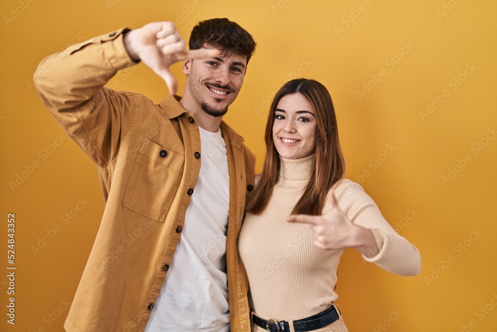 Fototapeta premium Young hispanic couple standing over yellow background smiling making frame with hands and fingers with happy face. creativity and photography concept.