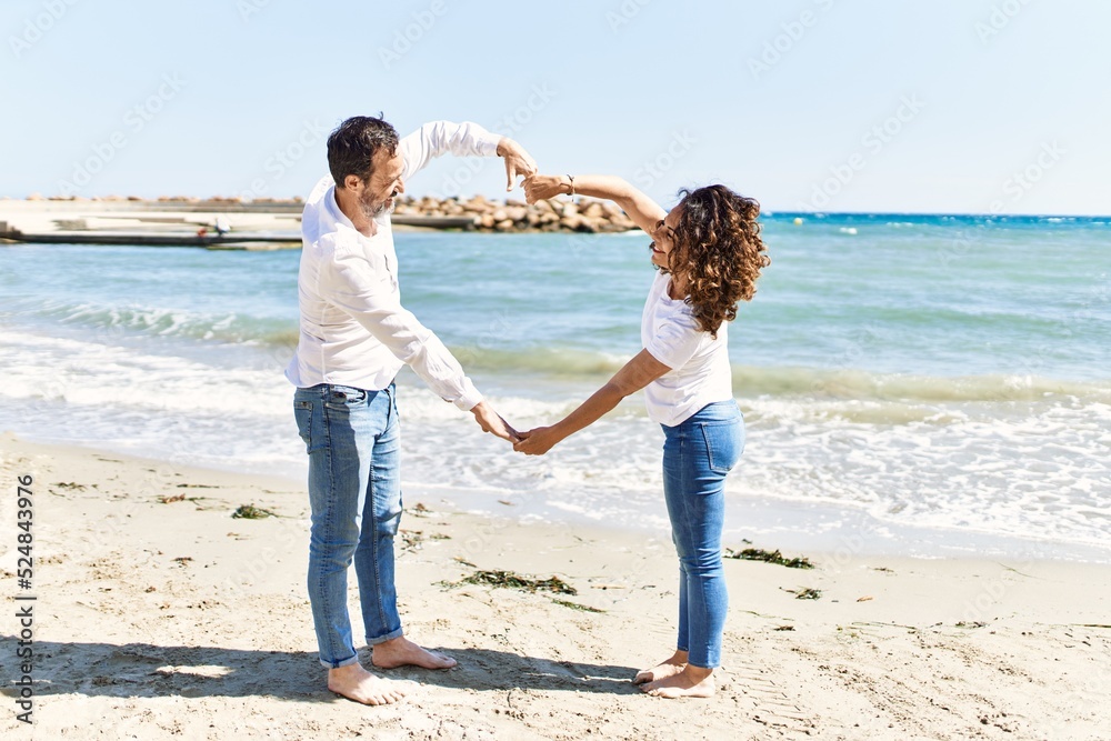 Middle age hispanic couple smiling happy doing heart gesture with arms at the beach.