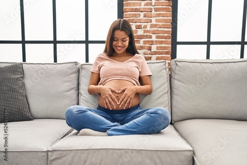 Young latin woman pregnant touching belly sitting on sofa at home