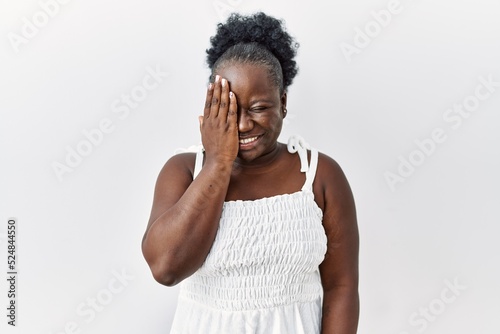 Young african woman standing over white isolated background covering one eye with hand, confident smile on face and surprise emotion.