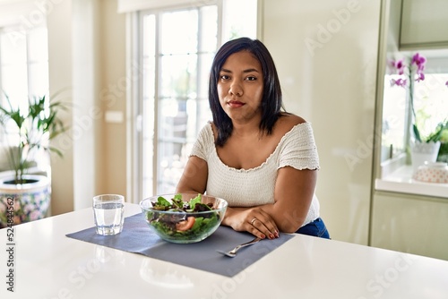 Young hispanic woman eating healthy salad at home relaxed with serious expression on face. simple and natural looking at the camera.