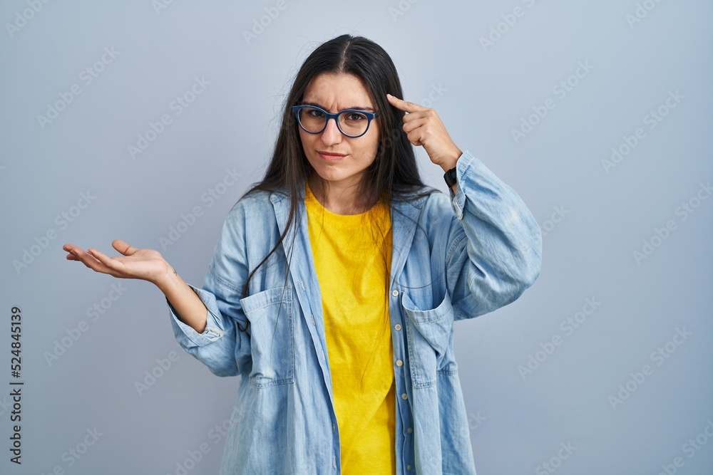 Young hispanic woman standing over blue background confused and annoyed with open palm showing copy space and pointing finger to forehead. think about it. Young hispanic woman standing over blue background confused and annoyed with open palm showing copy space and pointing finger to forehead. think about it.
